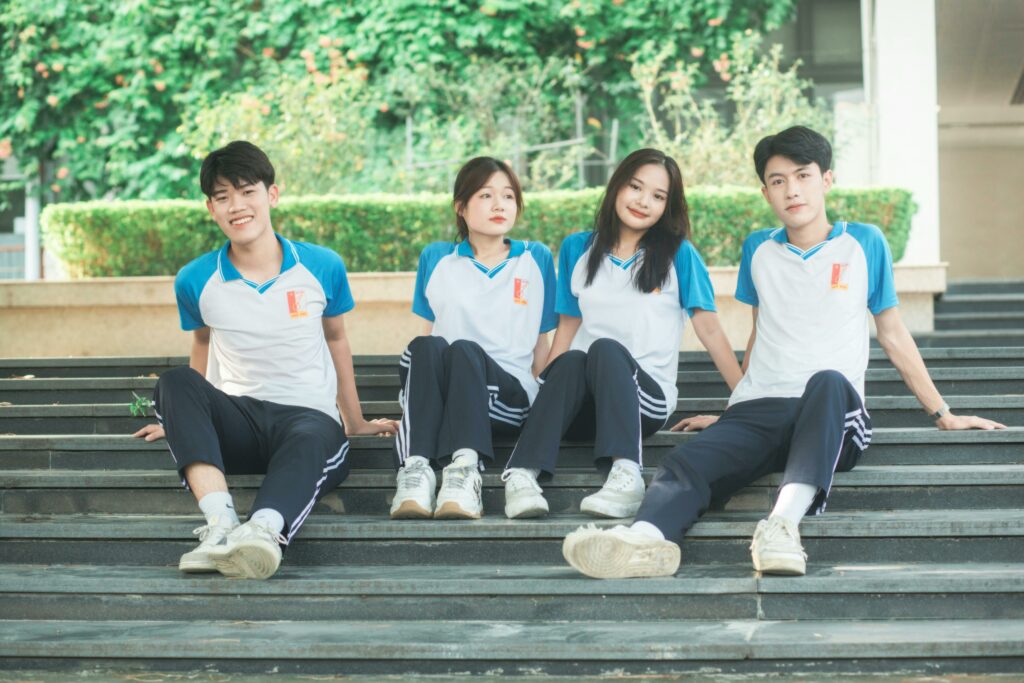 Four students wearing sports uniforms sitting on steps in a park area, looking relaxed and cheerful.