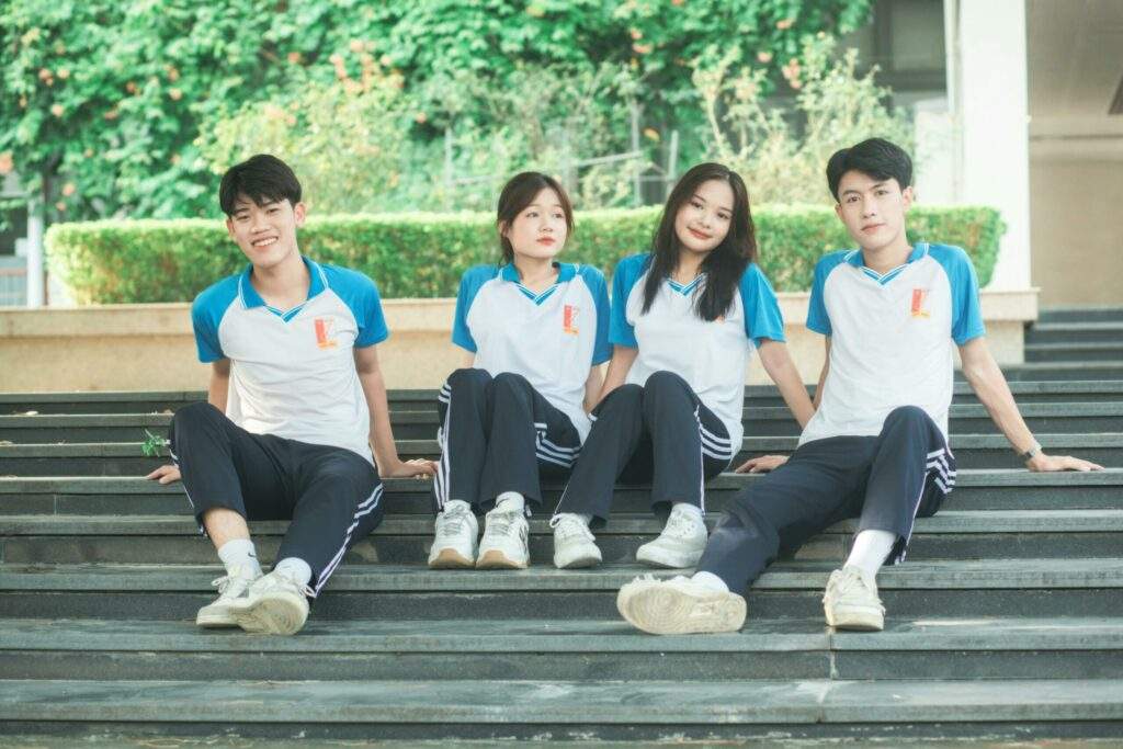 Four students wearing sports uniforms sitting on steps in a park area, looking relaxed and cheerful.