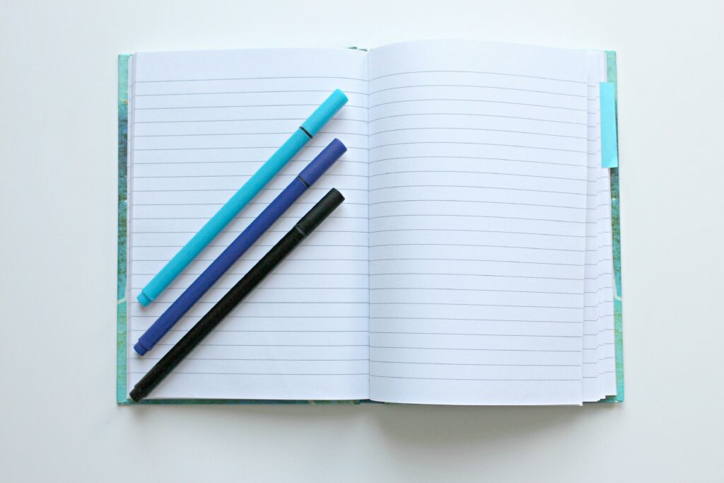 Open lined notebook with colorful pens on a white surface, perfect for note-taking and office tasks.
